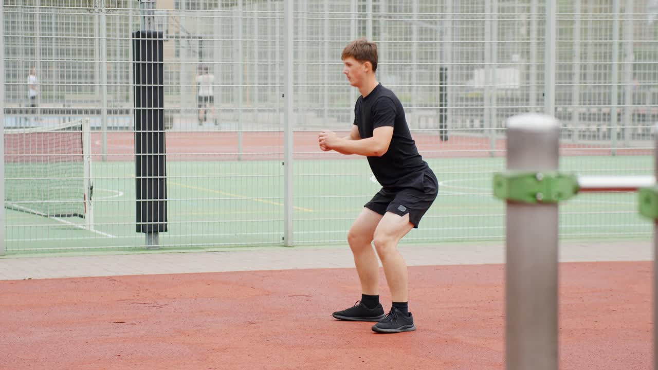 Un giovane uomo esegue squat a corpo libero all'aperto, impegnato in un allenamento di fitness su un campo sportivo, allenandosi per la forza e la salute fisica, telecamera statica, slow motion