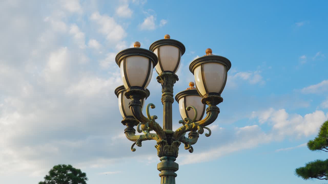 A low-angle close-up shot of a decorative, multi-headed vintage lamppost with ornate metalwork set against a bright, partly cloudy blue sky