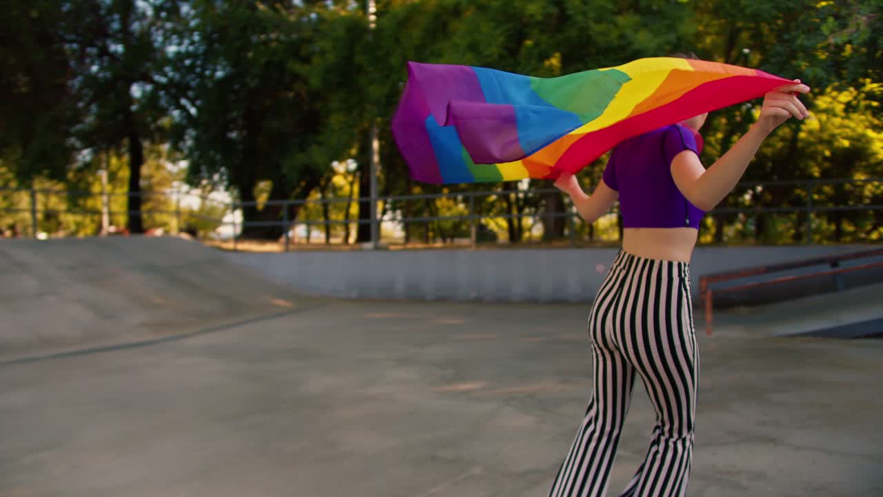 vista trasera de una chica con una camiseta púrpura y pantalones a rayas patinando en un parque de patinaje y sosteniendo una bandera lgbt ondeando