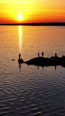 people standing on stones near the water in the evening. drone view on the natural background of the river and silhouettes of people at sunset. camera moves up back. vertical video