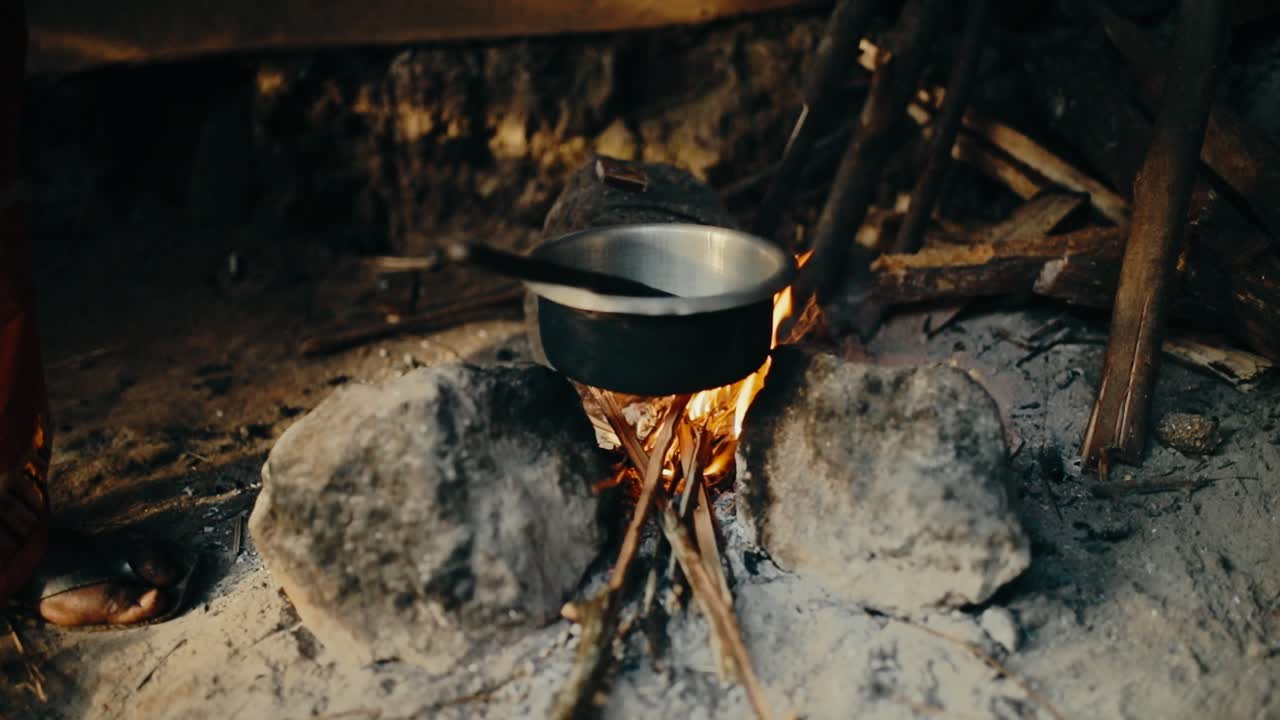 Feeding a wood fire under metal bowl to roast coffee beans - African person