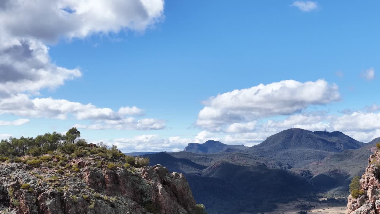 Camera slowly pans across a sunlit rocky outcrop with sparse vegetation, revealing expansive mountain ranges under a bright blue sky with scattered clouds