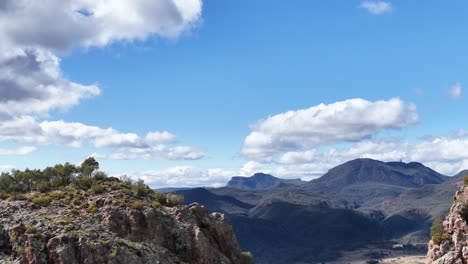 camera slowly pans across a sunlit rocky outcrop with sparse vegetation, revealing expansive mountain ranges under a bright blue sky with scattered clouds