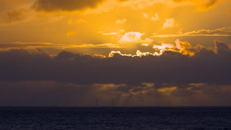 Dark-clouds-sweeping-by-along-horizon-with-spinning-wind-turbines-as-setting-sun-is-obscured-on-summer-evening