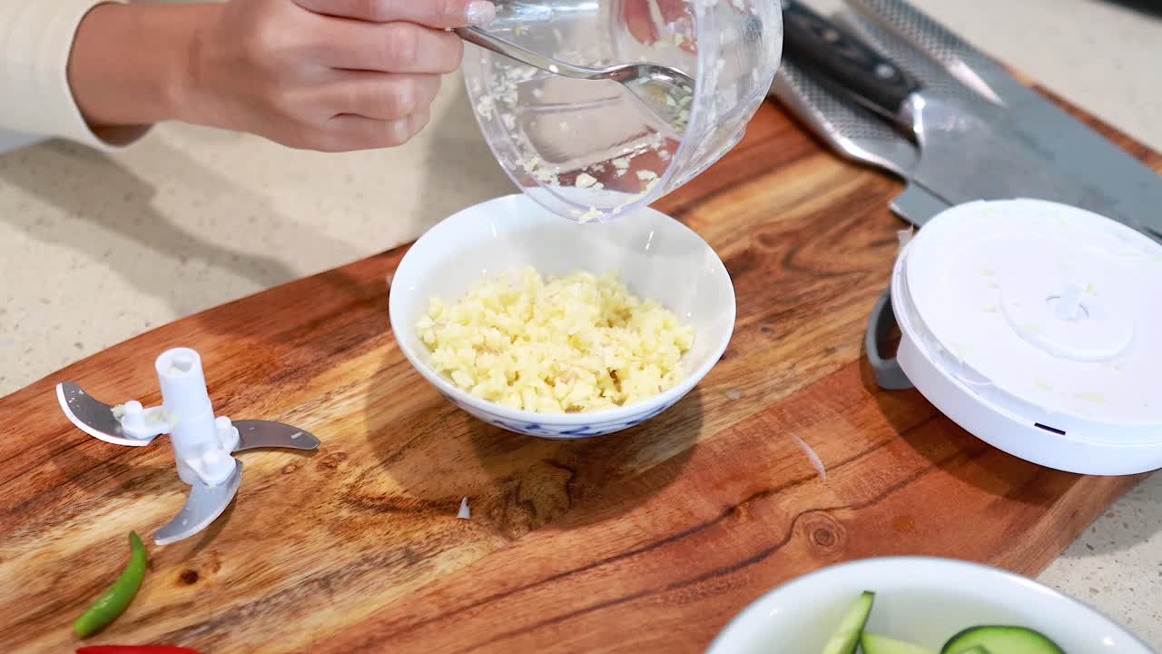 Hands pour minced garlic into a bowl on a wooden board. Bright lighting highlights the kitchen's clean, organized environment