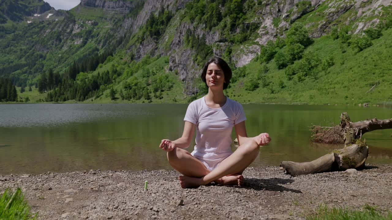 Woman meditating by a serene mountain lake surrounded by lush green hills on a sunny day