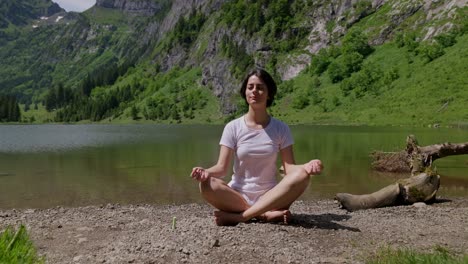 woman meditating by a serene mountain lake surrounded by lush green hills on a sunny day