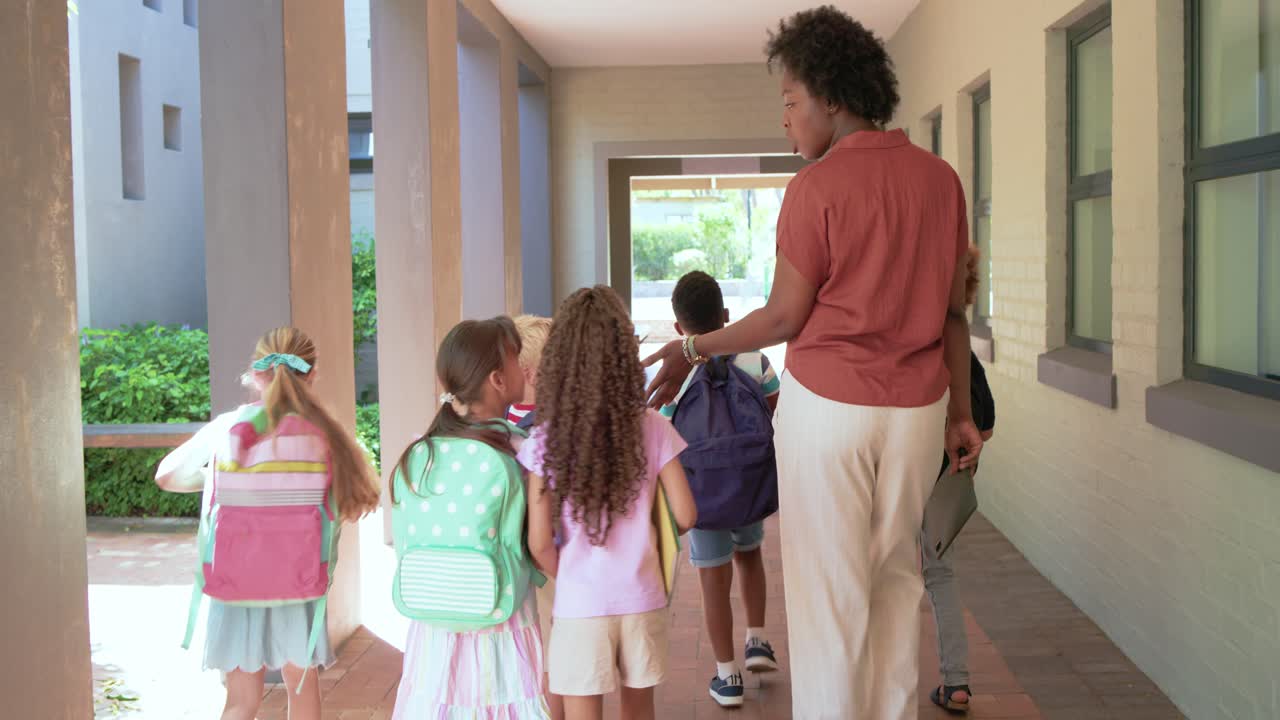 African American teacher guiding six youth, touching backpacks and holding tablet toward outdoors