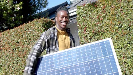 Retrato-De-Un-Hombre-Afroamericano-Feliz-Sosteniendo-Un-Panel-Fotovoltaico-Entrando-A-La-Propiedad,-Cámara-Lenta