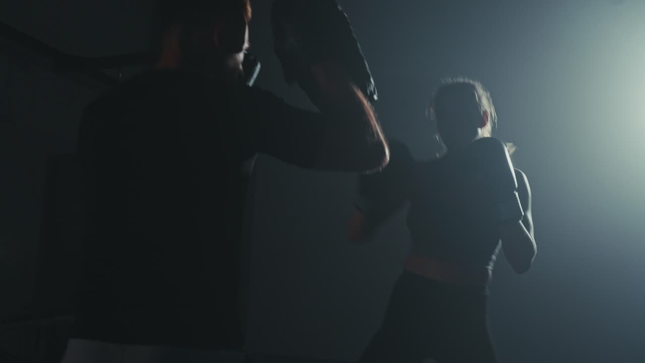 Kvinnlig boxare tränar med tränare i ett mörkt gym med dramatisk belysning