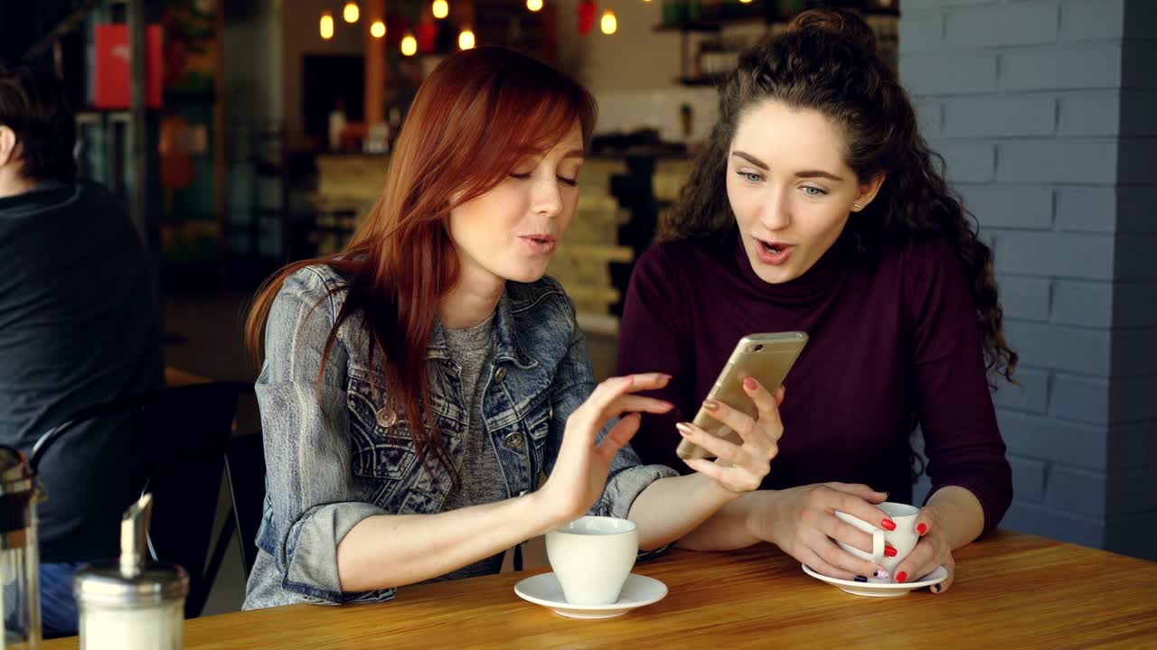 Pretty young girl is using smartphone and showing interesting photos to her female friend the discussing them while drinking coffee in nice modern cafe.