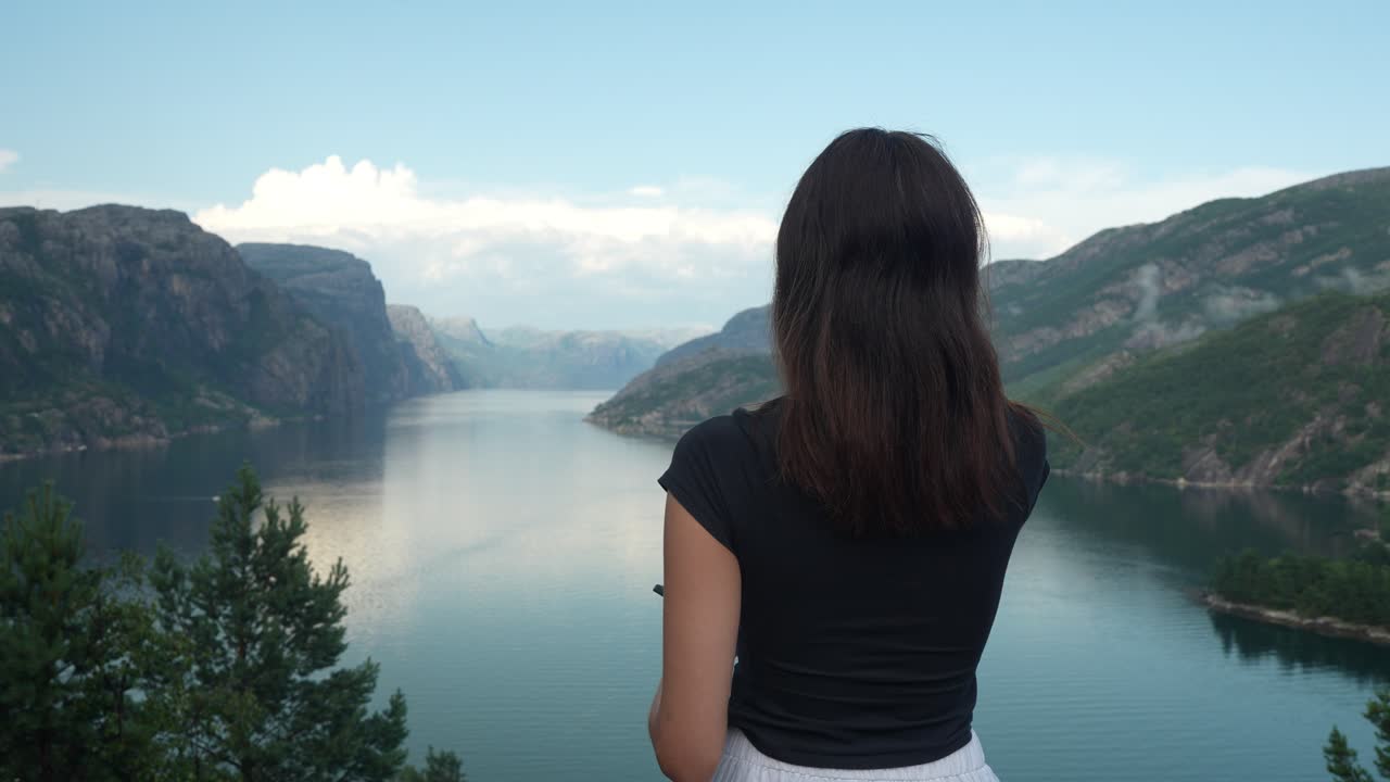 From a high vantage point, a woman with long dark hair stands by a fjord, gesturing towards the serene waters and rocky hills that define the breathtaking landscape around her