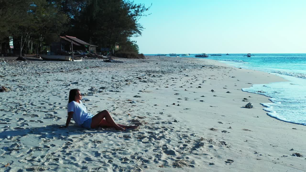 Thoughtful girl sitting on shiny white sand of exotic beach watching beautiful sea waves and ocean horizon touching bright blue sky in Bali