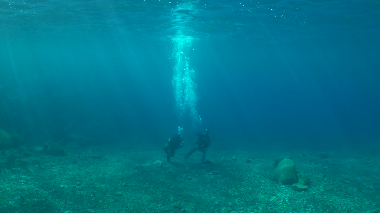 Two scuba divers talking and resting on the shallow seabed, with bubbles rising, in the underwater landscape