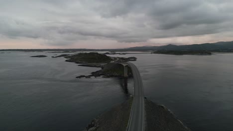 Movimiento-Lento-De-Vuelo-Sobre-El-Camión-Que-Pasa-Por-La-Carretera-Del-Océano-Atlántico-En-Noruega-Al-Atardecer-Con-Los-Faros-Encendidos-Y-La-Puesta-De-Sol-Y-Las-Montañas-Cubiertas-De-Nubes-En-El-Fondo