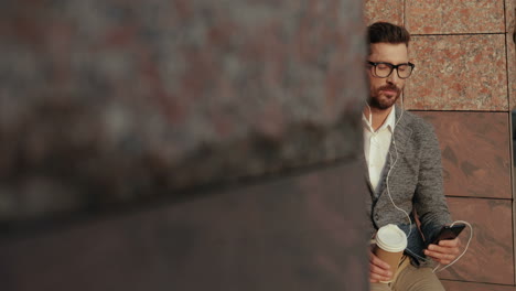 relaxed businessman drinking coffee and listening music on the smartphone via earphones while sitting on steps of a building in the city