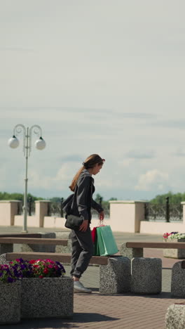 dama de ropa gris camina hacia un banco al aire libre con flores cerca, su ropa revoloteando en el viento, deja caer sus bolsas de compras, se sienta, y ajusta su posición cruzando la pierna