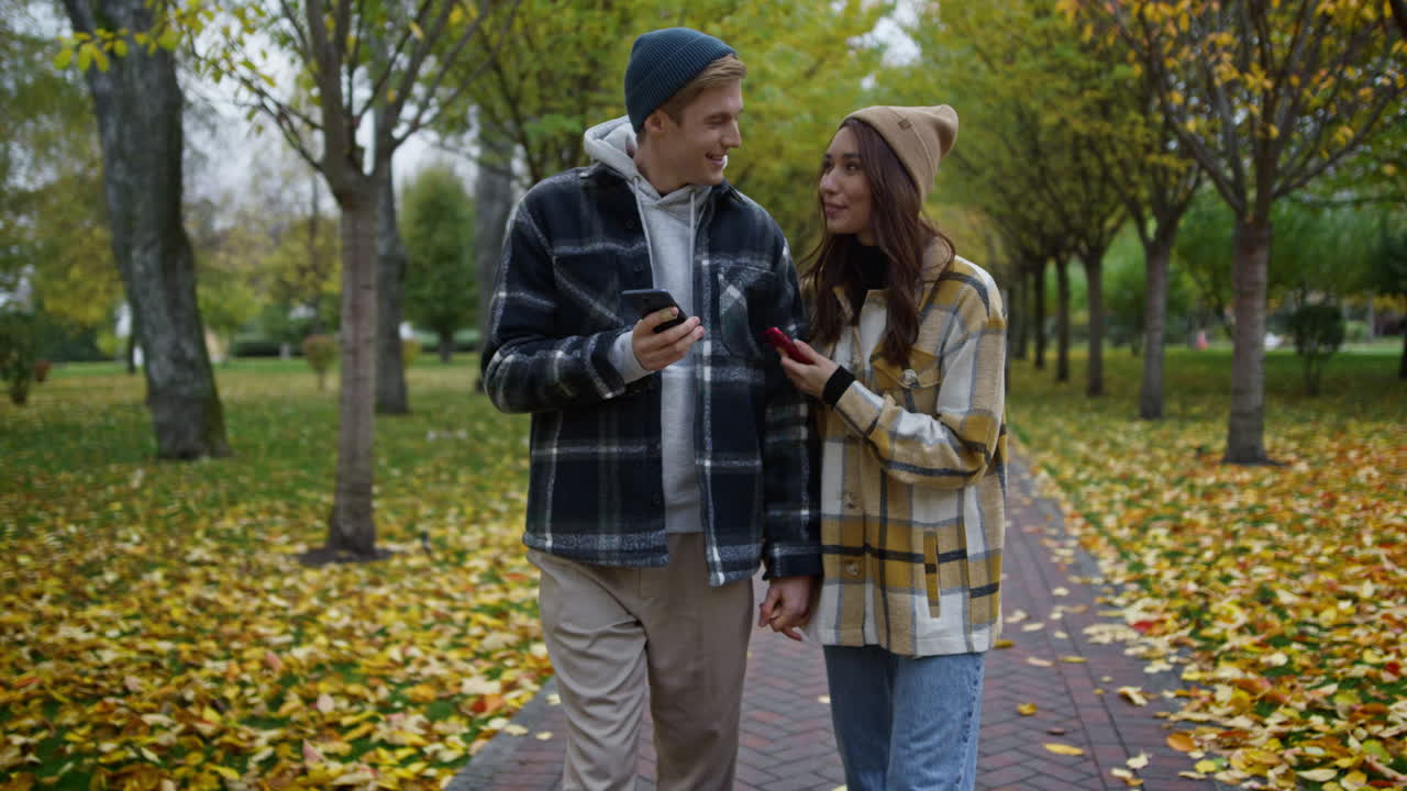 Giovane coppia sorridente che guarda gli smartphone in un parco autunnale