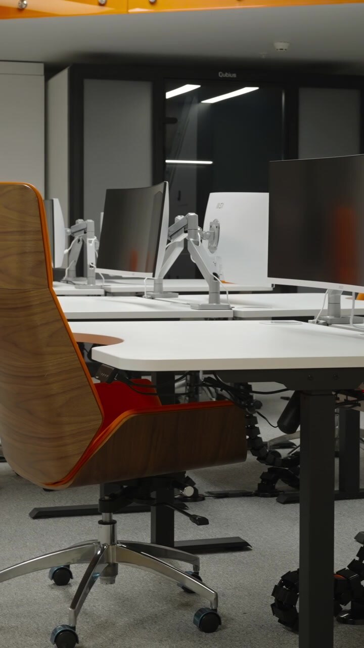 Interior de um local de trabalho contemporâneo e luminoso com mesas brancas, computadores de mesa, cadeiras laranja, divisória de vidro, escada e um banco verde em primeiro plano.