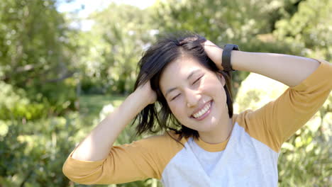 smiling asian woman in yellow shirt enjoying outdoors, hands in hair, wearing smartwatch