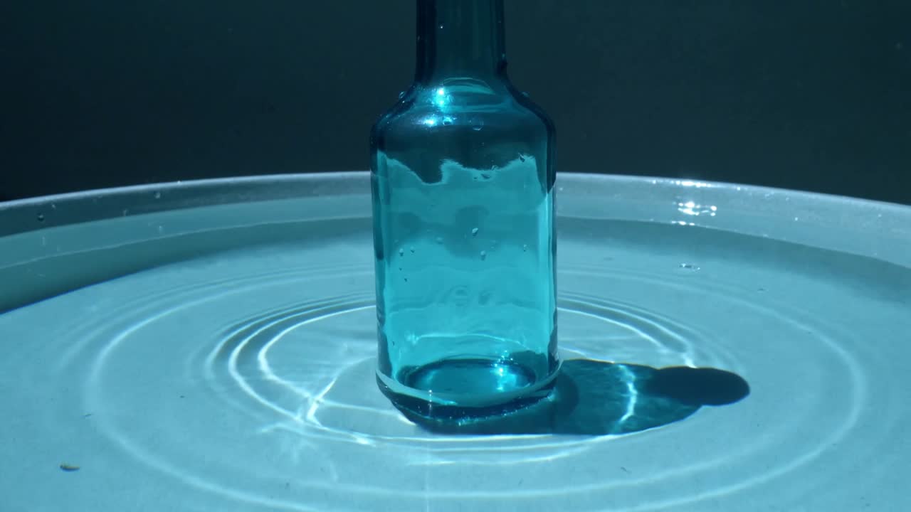 Riak awal dimulai dari botol kaca biru, menyebarkan gelombang ke seluruh nampan, dengan pantulan cahaya yang bergeser.