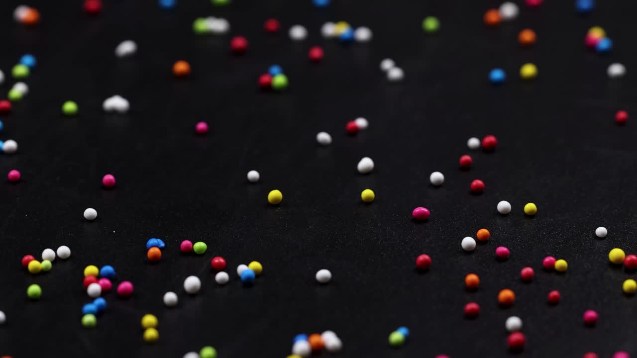 A variety of colorful sprinkles are scattered randomly across a dark background, creating a lively and playful pattern.