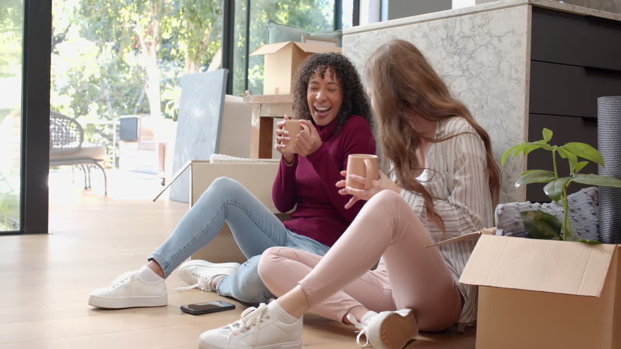 Relaxing on floor, lesbian couple drinking coffee and laughing while unpacking boxes
