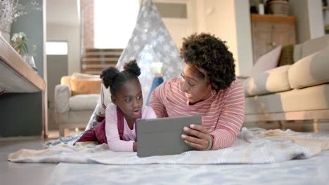 Happy-african-american-mother-and-daughter-lying-outside-tent-and-using-tablet-at-home,-slow-motion