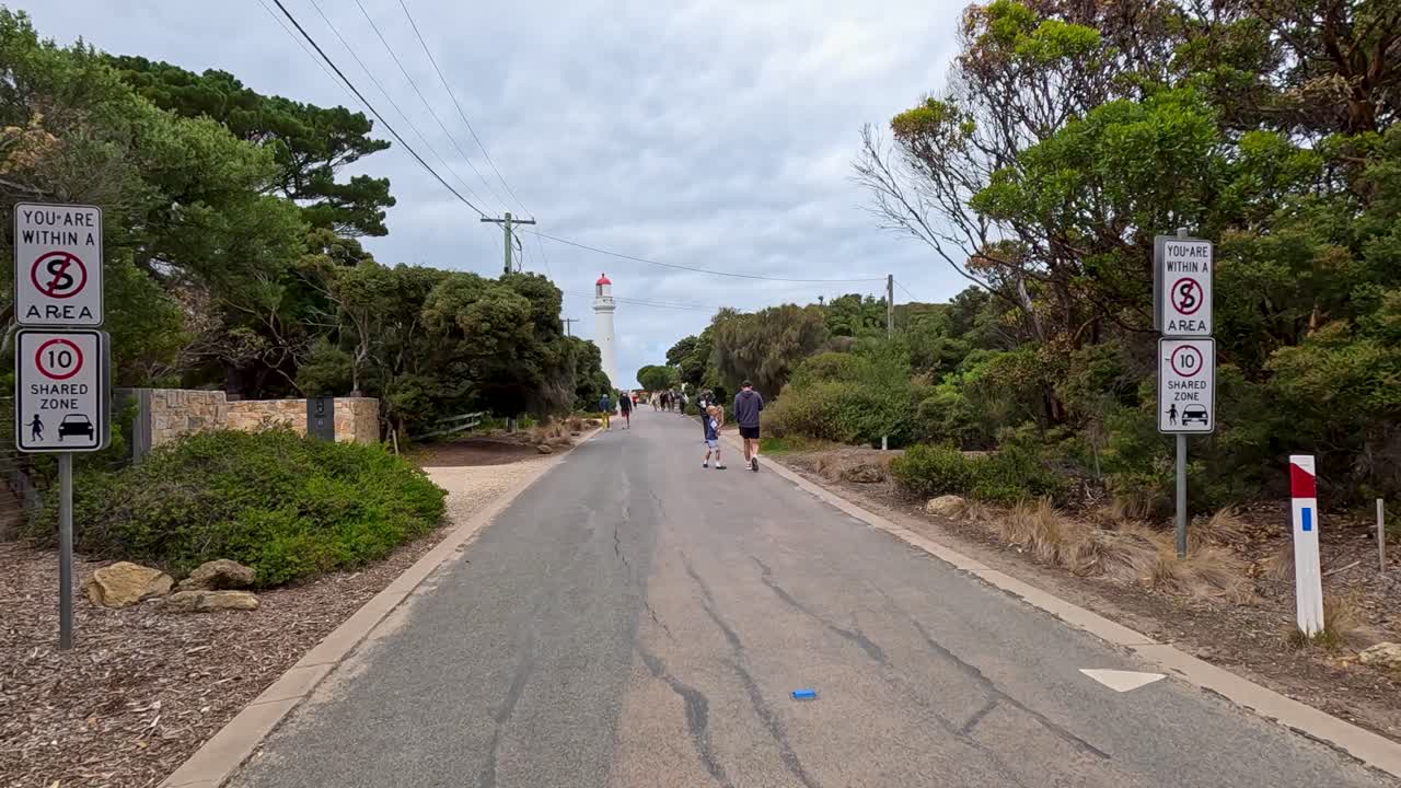 Several people walk down a quiet, tree-lined road under overcast skies near Bells Beach, with steady forward camera movement and natural daylight