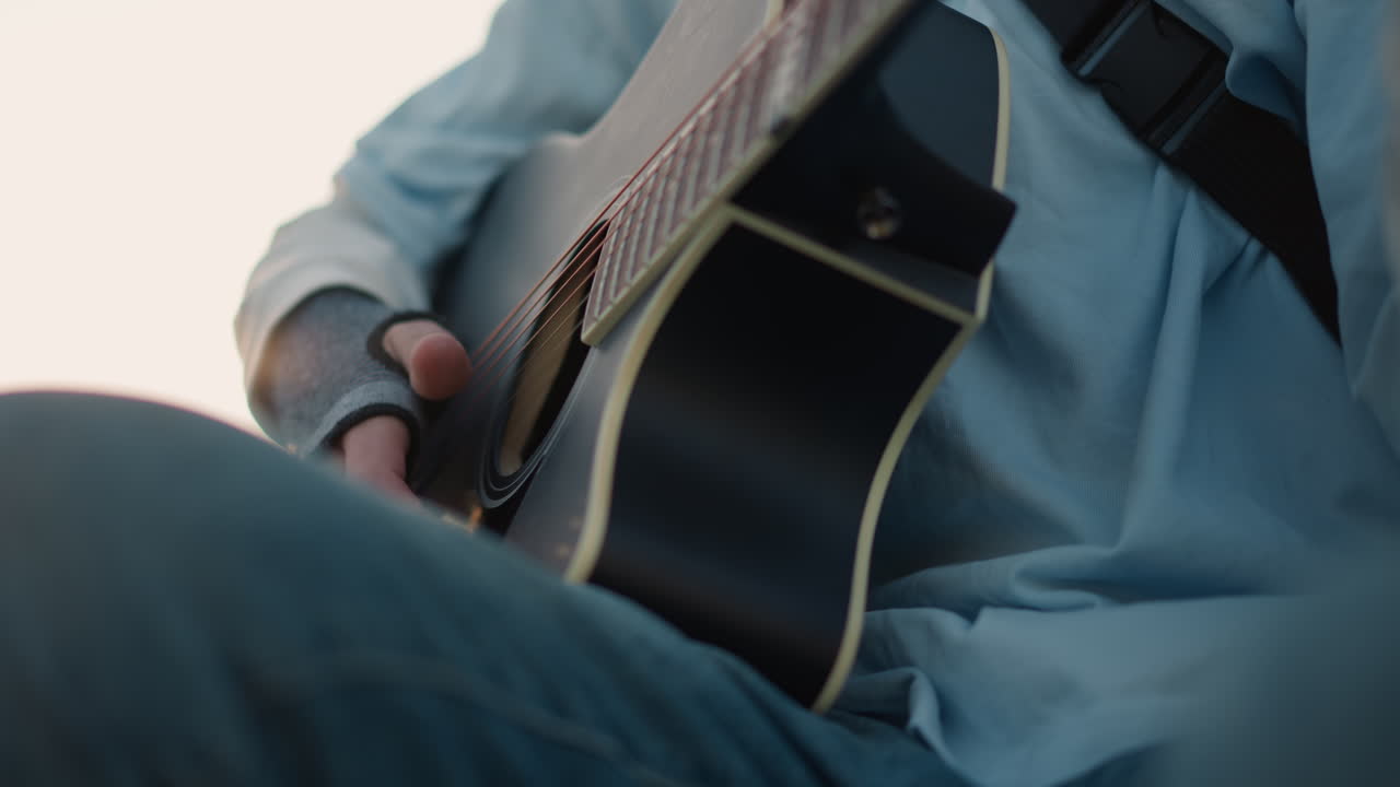 cantautore bianco che suona la chitarra acustica all'aperto durante l'ora d'oro, adolescente in maglione che pizzica accordi delicati, sfondo di alberi di pino, espressione contemplativa, profondità di campo cinematografica ridotta, slow motion
