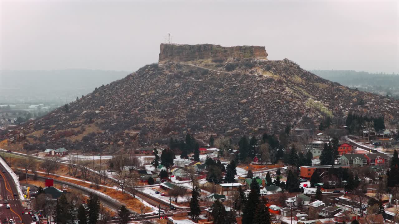 Veduta aerea con drone del centro di Castle Rock, Colorado, un punto di riferimento invernale, che mostra lo skyline della città e la mesa innevata del Front Range, con auto, edifici e quartieri in una fredda e nuvolosa mattina nevosa, mentre la neve cade su South Denver, ripresa con un movimento dinamico.
