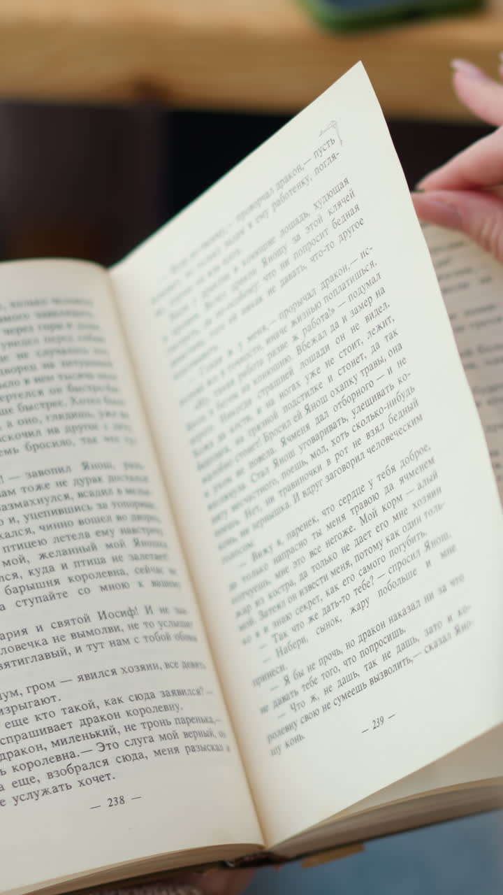 primer plano de la mano de la dama deslizándose sobre las páginas del libro mientras trata de volver a una nueva página mientras otra página se dobla hacia atrás, mesa de madera con teléfono visible en un fondo borroso