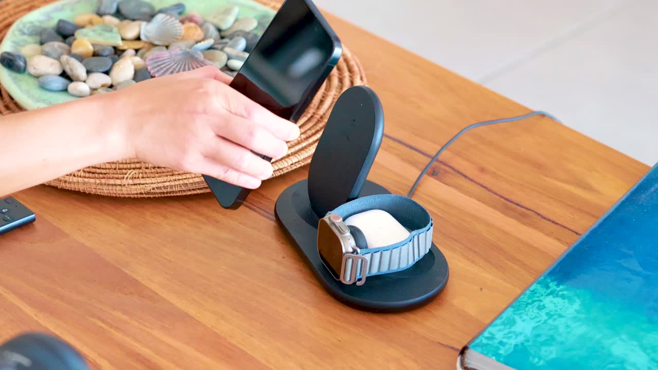 A person places a smartphone onto a wireless charging dock beside a smartwatch on a wooden table in a brightly lit indoor setting
