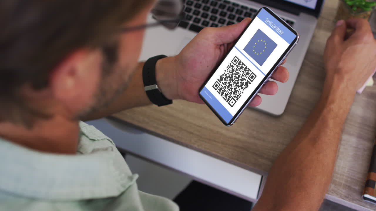 Man at desk holding smartphone with covid vaccination certificate, eu flag and qr code on screen