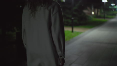 rear view woman in white coat walking alone at night on quiet path under gentle lamp glow, curly hair visible, benches lining walkway, capturing sense of calm solitude