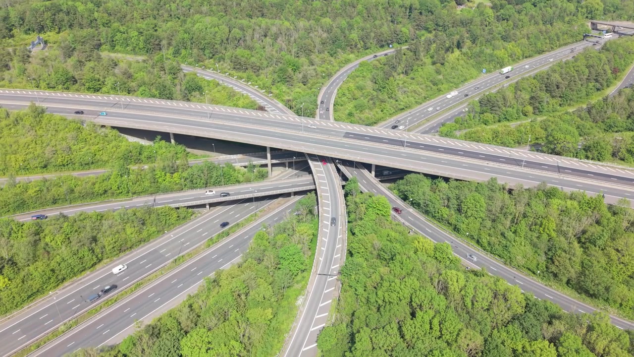 Aerial view of a complex highway interchange with multiple lanes and bridges. Vehicles are moving smoothly through the intersection surrounded by lush green forest during the day.
