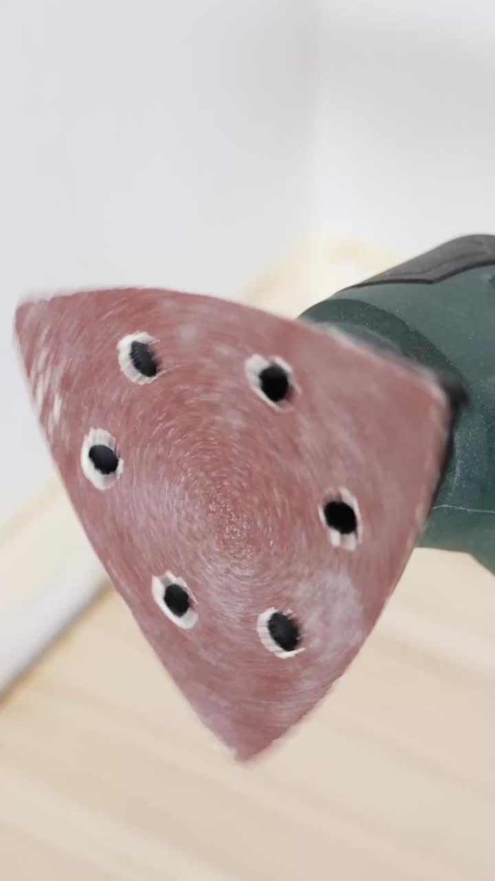 Vertical image shows a close-up of a triangular orbital sander tool with vibrating red sanding pad, ready for use on hardwood flooring during a surface preparation phase in a renovation project
