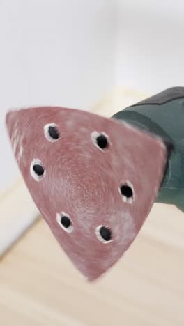 vertical image shows a close-up of a triangular orbital sander tool with vibrating red sanding pad, ready for use on hardwood flooring during a surface preparation phase in a renovation project