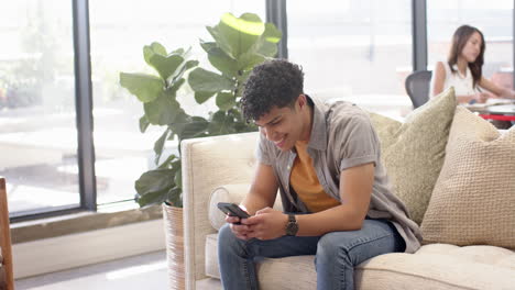Sitting on couch, young man using smartphone, woman working in background, at office