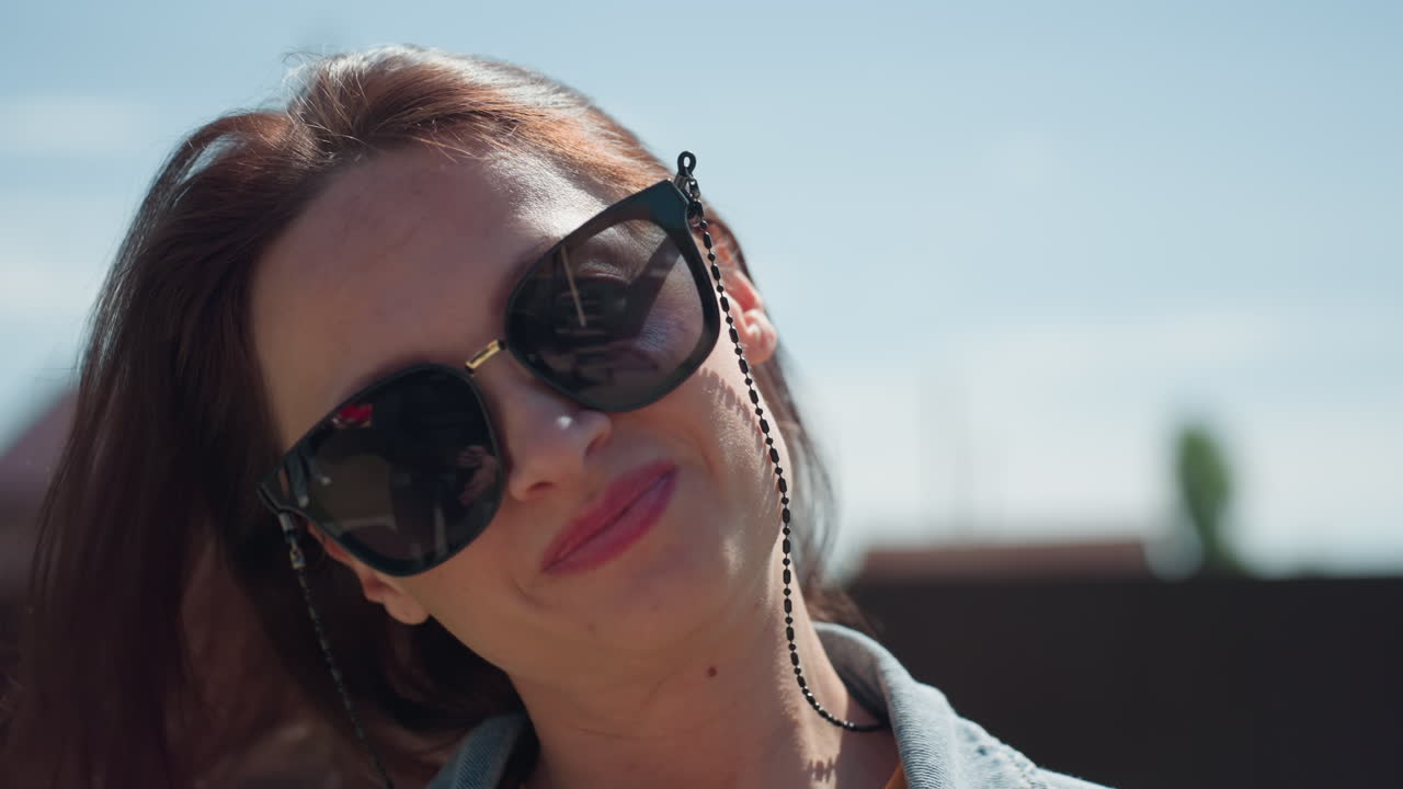 Confident young woman with long brown hair wearing orange shirt and jeans poses in front of camera, gently adjusting hair while smiling on bright sunny day with soft blurred trees and rooftops behind