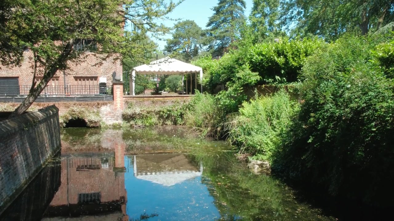 Ampia vista panoramica dello stagno a Well Hall Park con i riflessi dell'edificio storico, del cielo blu e degli alberi circostanti che creano un paesaggio estivo pacifico a Eltham, Londra.