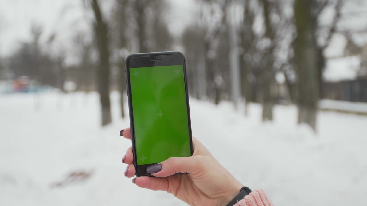 una mujer joven escribe y desliza en su teléfono inteligente en el parque de invierno. teléfono celular con pantalla de teclas cromadas - pantalla verde. cerrar. seguimiento de movimiento.