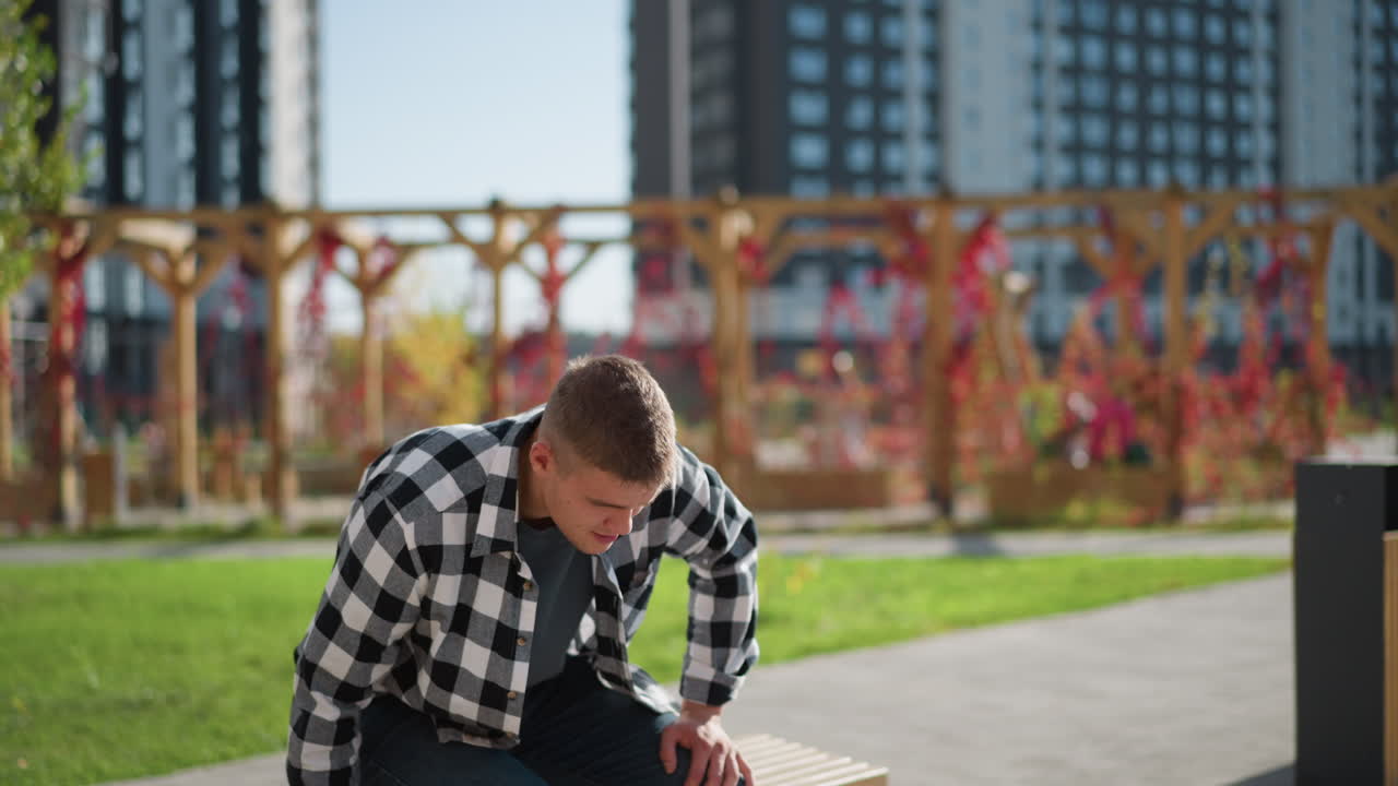 young student in checkered shirt bends forward to sit on wooden bench in bright urban park with residential buildings in background and gently pulls medicine from pocket under soft outdoor light