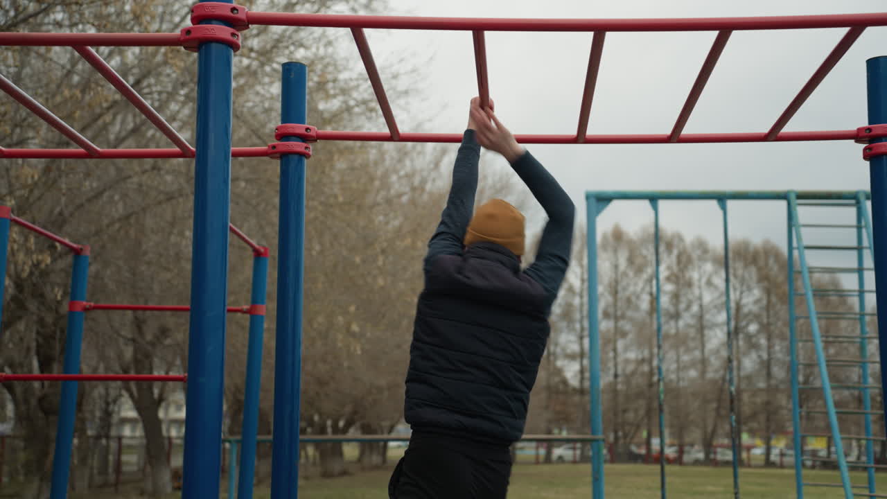 Close-up of a coach holding onto a red iron bar attached to blue poles, turning his body as he hangs and eventually drops down with a bounce