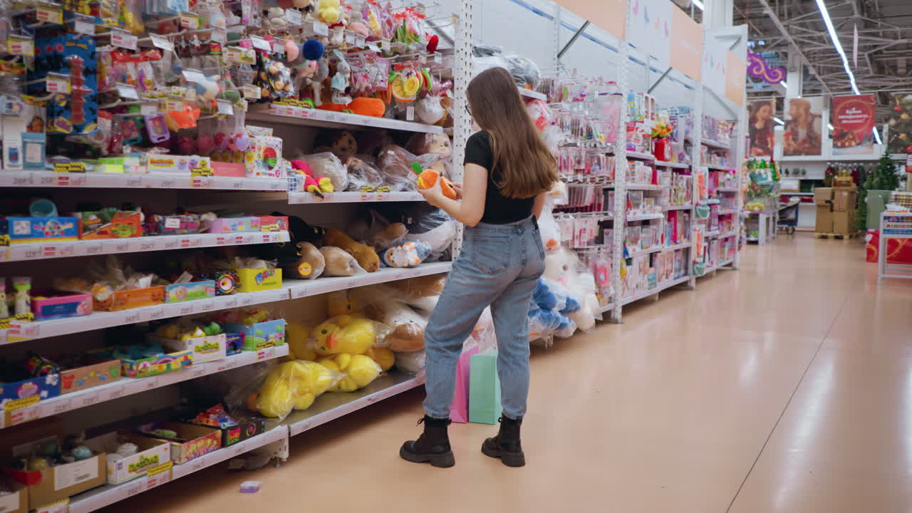 joven con camiseta negra sostiene e inspecciona un vibrante bolso de peluche naranja con detalles verdes en un centro comercial bien iluminado, examina cuidadosamente el artículo mientras navega por los estantes llenos de juguetes coloridos