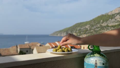 Hand-taking-olive-from-plate-on-balcony,-seaside-town-view,-Komiza,-Vis,-Croatia