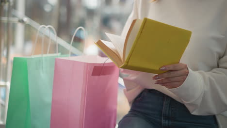 close-up of a woman seated indoors, flipping through a yellow-covered book with colorful shopping bags by her side, capturing a casual and relaxed moment in a bright modern shopping mall setting