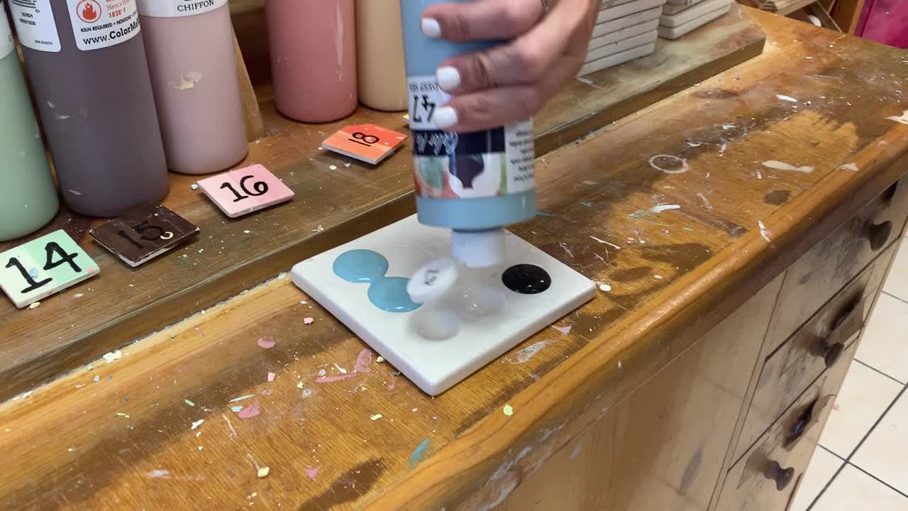 Hand squeezing a blue paint bottle and choosing colors on a white paint palette at "Color Me Mine Art" and Pottery studio in Saratoga, California.