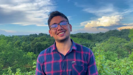 A-man-with-black-hair-and-glasses-wears-a-chemise-and-laughs-with-a-lovely,-happy-expression-while-standing-in-a-green-forest-with-stunning-clouds-in-the-background