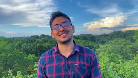un hombre con cabello negro y anteojos usa una camisa y se ríe con una expresión encantadora y feliz mientras está de pie en un bosque verde con impresionantes nubes en el fondo